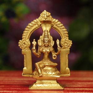 Peut inclure: Statue en laiton doré représentant une divinité assise, ornée de détails complexes. La figure est encadrée par une structure arquée avec des motifs de serpent. La statue repose sur une base carrée, mettant en valeur l'artisanat traditionnel.