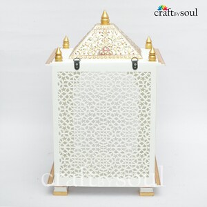 以下が含まれることがあります： 前面パネルに幾何学的な切り絵模様が施された白い装飾ランタン。上部には金色のアクセントと花の模様が施されたピラミッド型。ランタンは、金色のトリムが付いた長方形のベースで支えられています。