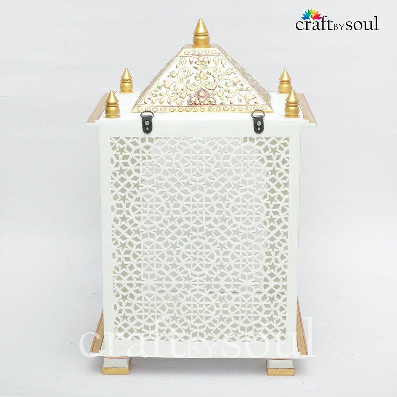 以下が含まれることがあります： 前面パネルに幾何学的な切り絵模様が施された白い装飾ランタン。上部には金色のアクセントと花の模様が施されたピラミッド型。ランタンは、金色のトリムが付いた長方形のベースで支えられています。