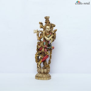 May include: A detailed brass statue of a deity playing a flute. The figure is adorned with intricate details, including a crown, jewelry, and a red and gold garment. The statue stands on a decorative base and is set against a white background.