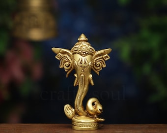 May include: A gold-colored brass statue of the Hindu deity Ganesha. The statue features intricate details, including elephant head, large ears, and multiple arms. The statue is on a wooden surface with a blurred background. The logo "craftbysoul" is in the top right corner.