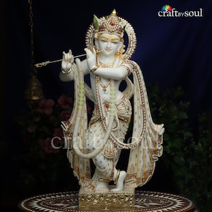 Puede incluir: Estatua blanca y dorada de una deidad tocando la flauta. La estatua está adornada con detalles intrincados, incluyendo una corona, joyas y vestiduras fluidas. La figura está arrodillada sobre una base decorativa, con un fondo azul oscuro.