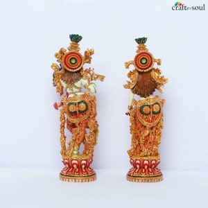 May include: Two ornate statues of a deity, one facing forward and the other backward. The statues are painted in vibrant colors, including orange, gold, green, and red. The deity is depicted with a flute and elaborate headdress, standing on a detailed base.