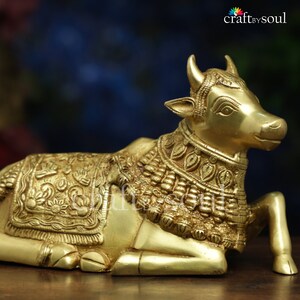 May include: A golden brass sculpture of a reclining cow, adorned with intricate floral and leaf patterns. The cow has detailed facial features and is wearing decorative neck and body ornaments. The sculpture is a religious or decorative item.