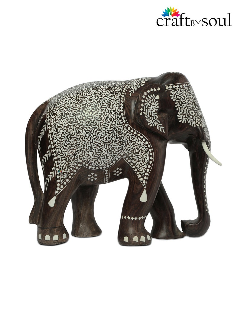 Puede incluir: Una figura de elefante de madera marr&oacute;n oscuro con intrincados patrones de incrustaciones florales blancas. El elefante tiene colmillos de color marfil y est&aacute; de pie sobre cuatro patas. El logotipo "craft by soul" est&aacute; en la esquina superior derecha.