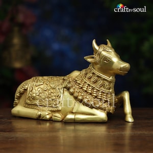 May include: A golden brass sculpture of a reclining cow, adorned with intricate floral and geometric patterns on its blanket and collar. The sculpture has small horns and is set on a wooden surface. The logo "craftbysoul" is in the top right corner.