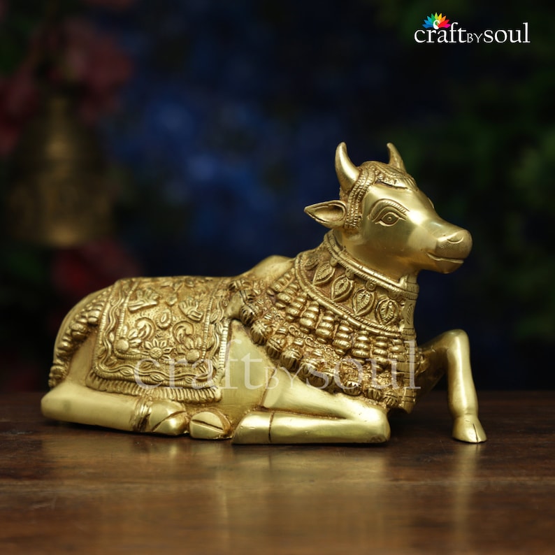 May include: A golden brass sculpture of a reclining cow, adorned with intricate floral and geometric patterns on its blanket and collar. The sculpture has small horns and is set on a wooden surface. The logo "craftbysoul" is in the top right corner.