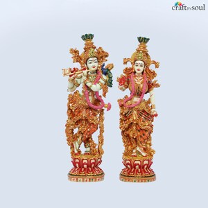 May include: Two ornate statues of deities, one playing a flute and the other holding a flower. Both figures are adorned with elaborate gold and red detailing, floral garlands, and headdresses. The statues stand on decorative red and gold bases, with a light blue background.