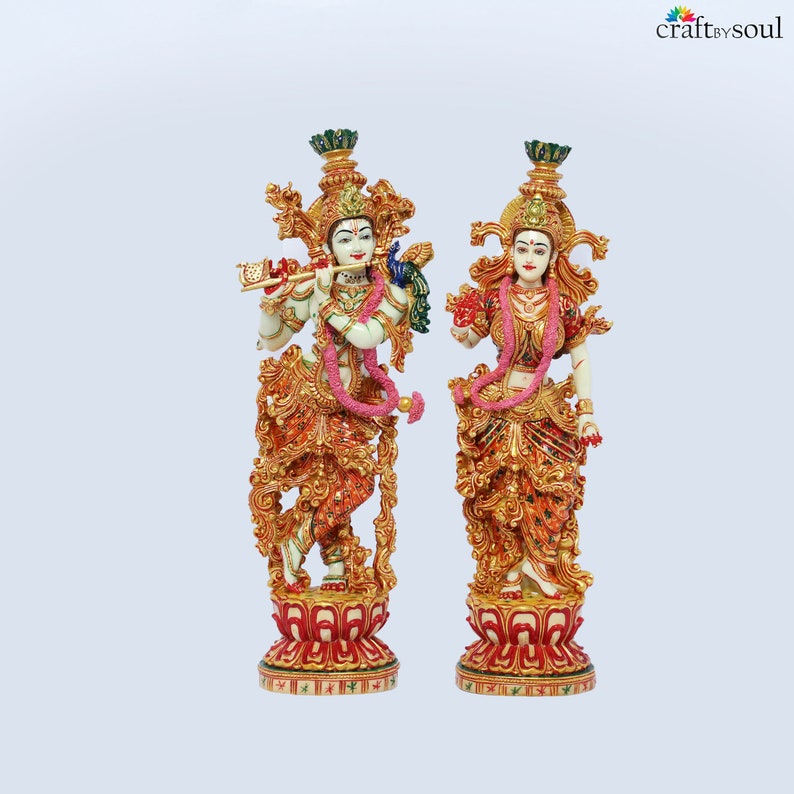 May include: Two ornate statues of deities, one playing a flute and the other holding a flower. Both figures are adorned with elaborate gold and red detailing, floral garlands, and headdresses. The statues stand on decorative red and gold bases, with a light blue background.