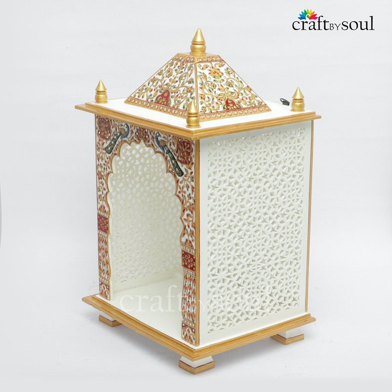 以下が含まれることがあります： 側面には複雑な切り絵模様が施された、白い木製の装飾寺院。 金色のトリムとアクセントが特徴で、屋根とアーチ型の入り口の周りには、詳細でカラフルなデザインが施されています。 寺院は小さな正方形の足の上にあります。