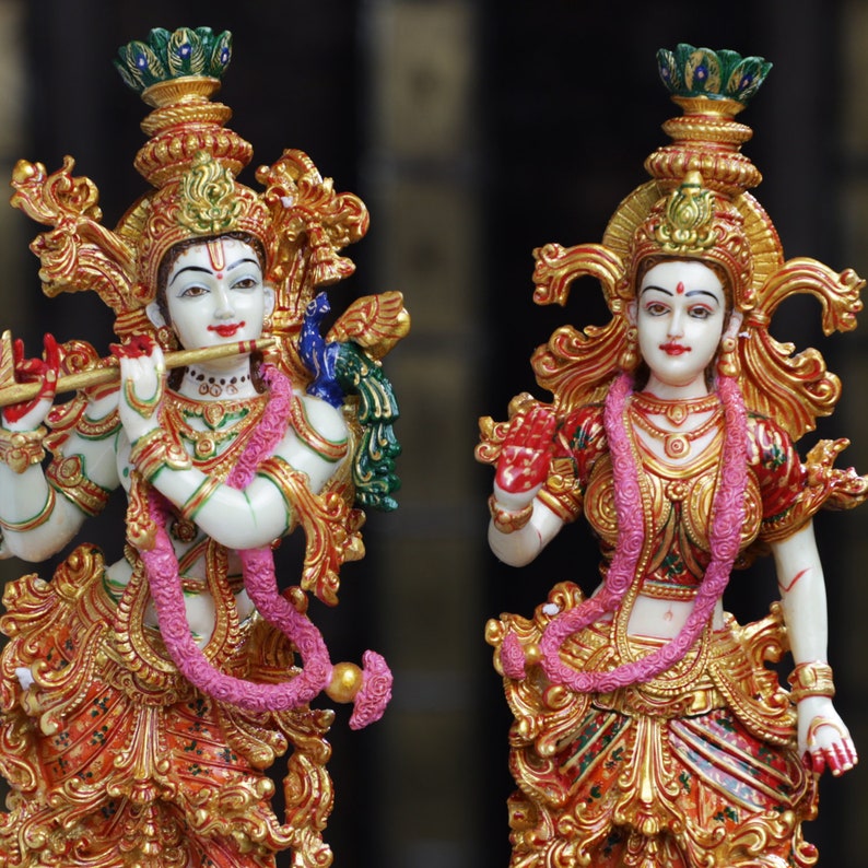 May include: Two ornate statues of Hindu deities. The figures are adorned with gold-colored embellishments, intricate detailing, and colorful accents. One figure holds a flute, and both wear pink garlands. The statues have a serene expression and are set against a dark background.