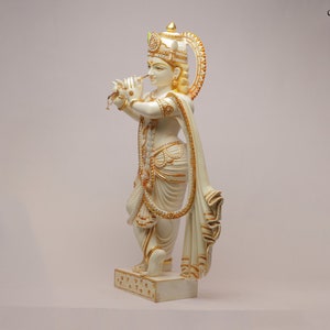 Puede incluir: Estatua de color crema de una figura tocando una flauta. La figura est&aacute; adornada con detalles dorados en la corona, las joyas y la ropa. La estatua est&aacute; sobre una base rectangular con un patr&oacute;n decorativo. El fondo es de un color claro s&oacute;lido.