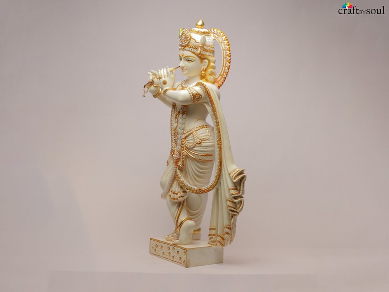 Puede incluir: Estatua de color crema de una figura tocando una flauta. La figura est&aacute; adornada con detalles dorados en la corona, las joyas y la ropa. La estatua est&aacute; sobre una base rectangular con un patr&oacute;n decorativo. El fondo es de un color claro s&oacute;lido.