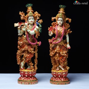 May include: Two ornate statues of Hindu deities. One figure plays a flute, adorned with a peacock. The other figure has a hand raised in blessing. Both statues are in gold, red, and white, with floral garlands and lotus bases.