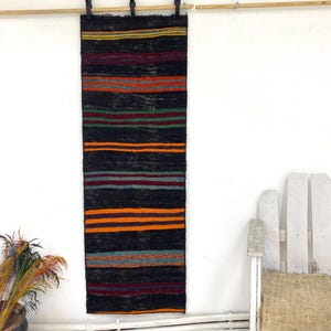 May include: A rectangular, woven wall hanging with horizontal stripes in black, orange, red, green, and blue. The rug is suspended from a wooden rod. The wall is white, and a white chair is visible on the right.