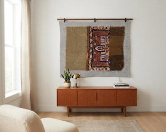May include: A rectangular wall hanging featuring a gray felted backing with a central panel of stitched fabric fragments in olive green, brown, and a patterned section with geometric motifs in red, blue, and cream, suspended from a wooden rod.