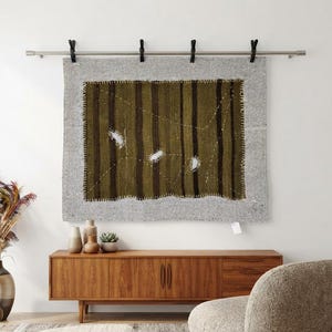 Peut inclure: Une tenture murale rectangulaire composée d'un panneau central vert olive à rayures verticales marron foncé avec des accents cousus blancs, bordé d'un tissu texturé gris clair. L'ensemble est suspendu à une tringle métallique argentée par quatre pinces noires.