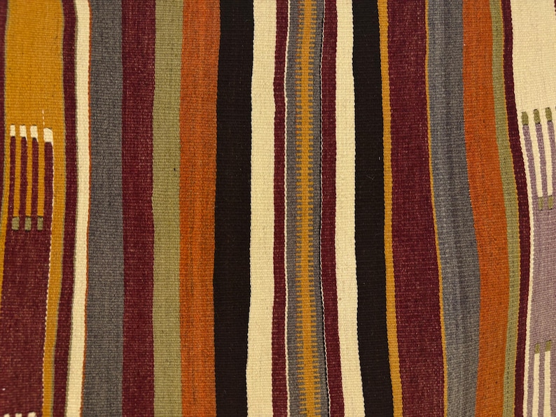 May include: A woven textile featuring a repeating vertical striped pattern in shades of deep burgundy, burnt orange, mustard yellow, charcoal gray, black, and off-white. The fabric displays a textured weave with small geometric accents.