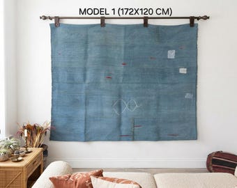 Op de afbeelding: Een groot rechthoekig blauw wandkleed met subtiele horizontale lijnen, kleine rode geborduurde accenten, witte geometrische vormen en lichtgekleurde lapjes. Het textiel hangt aan een donkerbruine roede met leren riemen.