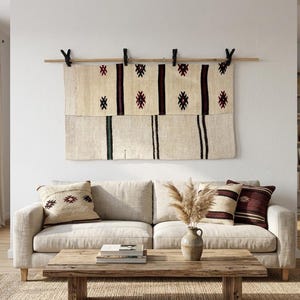 May include: A rectangular woven wall hanging featuring a cream-coloured base with vertical dark brown stripes and small geometric diamond motifs. The textile is suspended from a wooden rod by four black clips, displaying a textured, hand-loomed appearance.