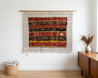 Pode incluir: Uma tapeçaria retangular com um tecido central apresentando faixas horizontais em vermelho, preto e amarelo, decoradas com padrões geométricos. Está montada numa barra de madeira sobre um fundo de tecido cinzento claro texturizado.