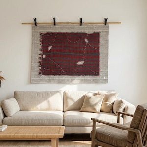 Peut inclure: Un tissage mural avec un motif rouge et gris est exposé au-dessus d'un canapé beige. La tenture murale est rectangulaire avec un bord effiloché et est suspendue à une tringle en bois avec des clips noirs. La pièce a une palette de couleurs neutres.