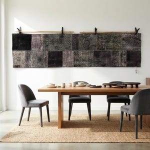 Op de afbeelding: Een lange, rechthoekige wandhanger bestaande uit donkere, getextureerde stoffen lappen in tinten zwart en antraciet, opgehangen aan twee houten roeden. Het stuk hangt boven een houten eettafel met vier grijze gestoffeerde stoelen.