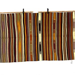 May include: A rectangular woven textile featuring a repeating vertical striped pattern in shades of deep burgundy, mustard yellow, olive green, dark brown, and cream. The fabric is suspended from two horizontal wooden rods using black clips.