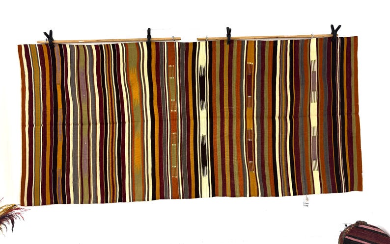 May include: A rectangular woven textile featuring a repeating vertical striped pattern in shades of deep burgundy, mustard yellow, olive green, dark brown, and cream. The fabric is suspended from two horizontal wooden rods using black clips.