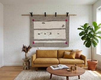 Puede incluir: Un tapiz rectangular con un centro de color crema con rayas horizontales negras y pequeños motivos geométricos negros, enmarcado por un borde gris ancho con cuatro borlas rosas en las esquinas, suspendido de una barra de madera con clips.