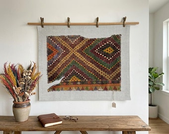 May include: A rectangular textile wall hanging featuring a geometric pattern in shades of red, green, yellow, and black, mounted on a light gray fabric backing and suspended from a wooden rod with four metal clips.