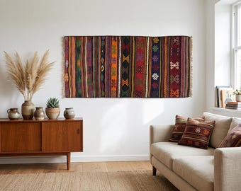 May include: A rectangular woven wall hanging featuring vertical stripes in shades of red, orange, green, purple, and black, decorated with repeating geometric patterns and finished with short fringe on the left and right edges.