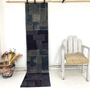 Puede incluir: Una alfombra rectangular larga hecha de cuadrados de tela grises y negros. La alfombra cuelga verticalmente contra una pared blanca. Una silla de madera blanca y una maceta de terracota con plantas secas también están en la imagen.