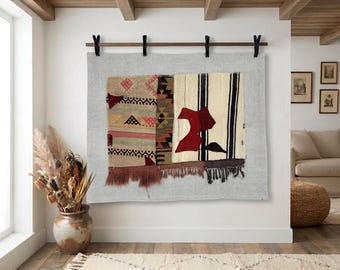 May include: A rectangular wall hanging featuring a light grey fabric background with a central patchwork panel of woven textile fragments. The panel displays geometric patterns, vertical stripes, and abstract shapes in red, black, tan, and cream, finished with a fringed bottom edge.