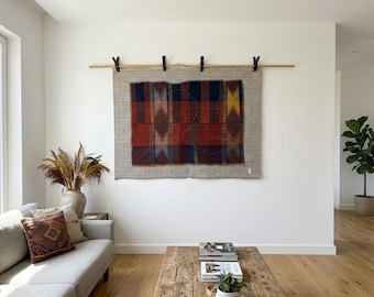May include: A rectangular textile wall hanging with a patchwork design in shades of red, blue, and yellow, framed by a neutral border. It is suspended from a wooden rod with black clips, adding a touch of rustic charm to the room.