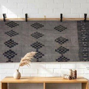 May include: A rectangular gray textile wall hanging featuring a repeating black diamond pattern and a vertical striped border, suspended from a light wood rod by six black metal clips against a white painted brick wall surface.