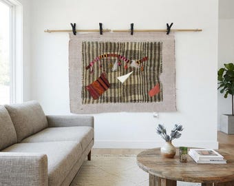 Puede incluir: Un tapiz rectangular con un patrón de rayas beige y marrón oscuro con bordes deshilachados. El tejido está decorado con un arco bordado curvo en rojo y amarillo, un parche triangular blanco y dos acentos de tela rectangulares más pequeños.