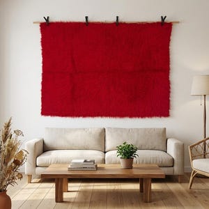 May include: A large, rectangular, vibrant red shag rug is mounted on a white wall. The rug is suspended from a wooden rod with black clips. The room features a neutral-toned sofa, wooden coffee table, and a rattan chair.