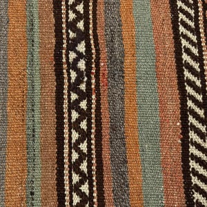 Op de afbeelding: Een geweven textiel met een reeks verticale strepen in tinten bruin, oranje en gedempt blauw. De stof vertoont een getextureerd oppervlak met afwisselende banen van effen kleuren en ingewikkelde donkerbruine zigzagpatronen op een cr&egrave;mekleurige achtergrond.