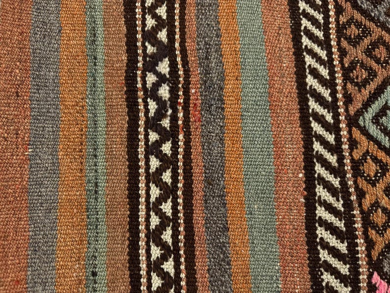 Op de afbeelding: Een geweven textiel met een reeks verticale strepen in tinten bruin, oranje en gedempt blauw. De stof vertoont een getextureerd oppervlak met afwisselende banen van effen kleuren en ingewikkelde donkerbruine zigzagpatronen op een cr&egrave;mekleurige achtergrond.