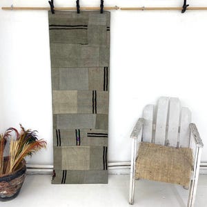 Può includere: Un tappeto rettangolare patchwork nei toni del grigio e del beige è appeso a una parete bianca. Strisce nere accentuano il design. Una sedia di legno bianca con seduta intrecciata e un vaso decorativo con piante essiccate sono anch'essi nell'immagine.