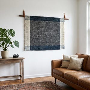 May include: A rectangular woven wall hanging featuring a dark navy blue centre with cream-coloured vertical side borders, suspended from a horizontal wooden rod with leather straps attached to the wall above a brown leather sofa and wooden console table.