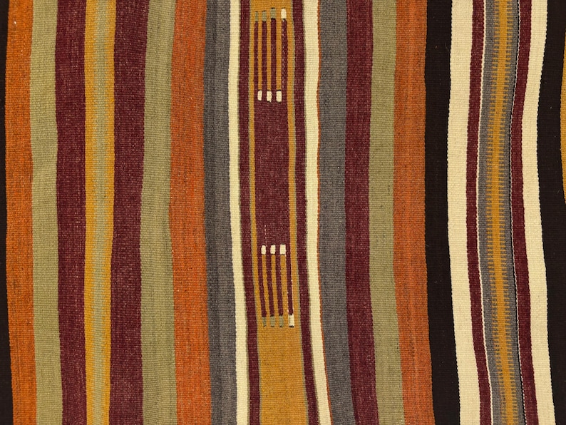 May include: A woven textile featuring vertical stripes in shades of burnt orange, deep maroon, olive green, mustard yellow, charcoal gray, and cream. The fabric displays a repeating pattern of small, light-colored rectangular stitch accents.