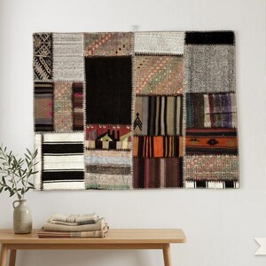 Puede incluir: Un tapiz de pared rectangular con un diseño de patchwork compuesto por varios segmentos de tela tejida en tonos negros, blancos, grises, naranjas y tierra. El textil muestra una mezcla de patrones geométricos, bloques sólidos y motivos a rayas.