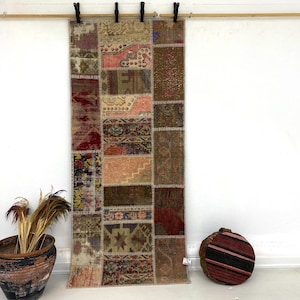 May include: A patchwork rug with a variety of colors, including red, brown, and beige, hangs from a wooden rod. The rug is made of rectangular pieces sewn together. A decorative pot with dried plants and a round, striped cushion are on the floor.