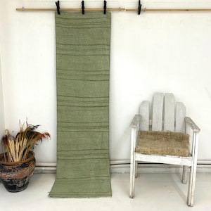 May include: A long, rectangular sage green table runner featuring thin, horizontal dark green stripes spaced at irregular intervals along its length. The fabric has a visible, slightly textured weave and straight, finished edges on all sides.