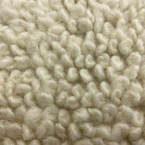 May include: Close-up of a cream-colored, textured fabric. The material has a thick, looped pile, creating a soft, plush appearance. The fibers are tightly packed, giving the fabric a dense, warm look. The texture is reminiscent of a sheepskin or faux fur.