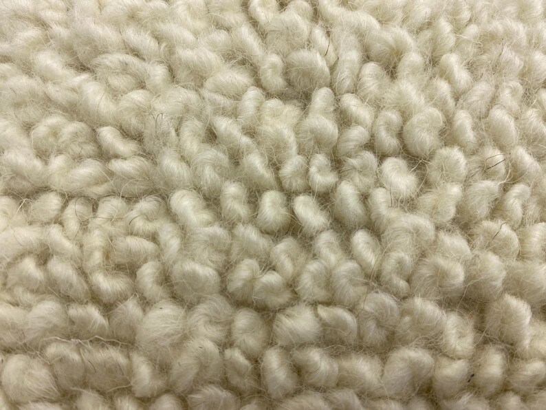 May include: Close-up of a cream-colored, textured fabric. The material has a thick, looped pile, creating a soft, plush appearance. The fibers are tightly packed, giving the fabric a dense, warm look. The texture is reminiscent of a sheepskin or faux fur.