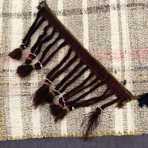 Puede incluir: Primer plano de una secci&oacute;n de un textil tejido con un patr&oacute;n de cuadros crema y marr&oacute;n. Flecos marrones oscuros con borlas est&aacute;n unidos al borde. El textil tiene un borde cosido amarillo y un fondo gris oscuro.