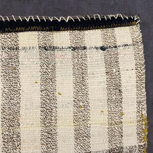 Puede incluir: Primer plano de una secci&oacute;n de un textil tejido con un patr&oacute;n de rayas en crema y marr&oacute;n. El borde est&aacute; acabado con un borde negro y costuras blancas. La esquina tiene un acento rojo y negro.